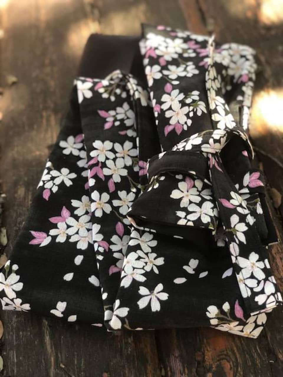 Tela negra con un estampado floral blanco y rosa, posiblemente una faja o cinturón. El patrón incluye flores de cerezo estilizadas. El fondo es una superficie de madera envejecida.