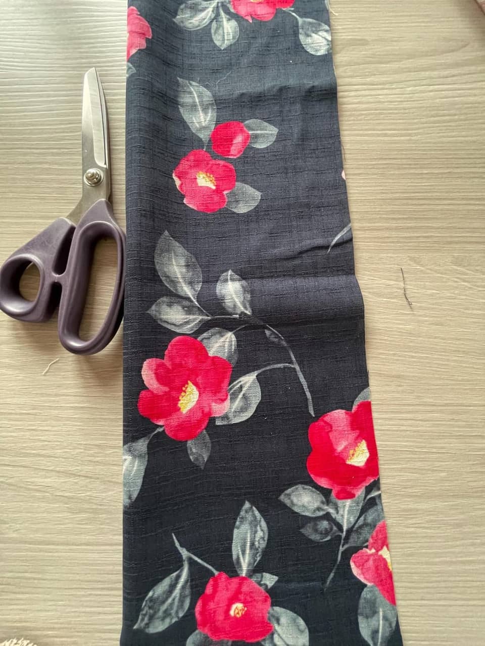 Unas tijeras con mango morado junto a un trozo de tela oscura. La tela está estampada con flores rojas y hojas grises. Las tijeras están abiertas, listas para cortar la tela.