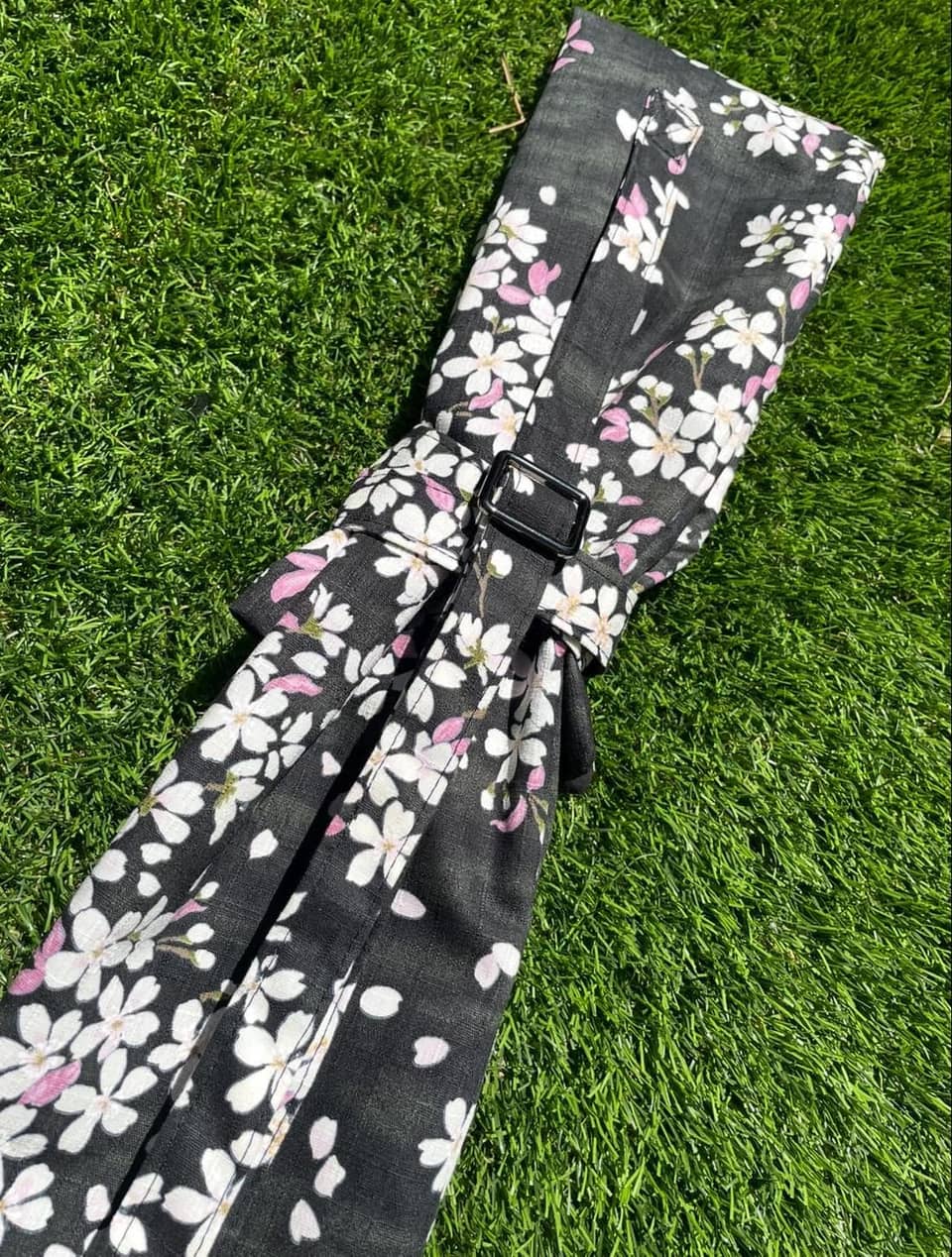 Un estuche de tela negro enrollable con un patrón floral de flores de cerezo en blanco y rosa. El estuche incluye una correa negra ajustable con una hebilla rectangular de plástico para asegurar la tela enrollada, con un estampado botánico.