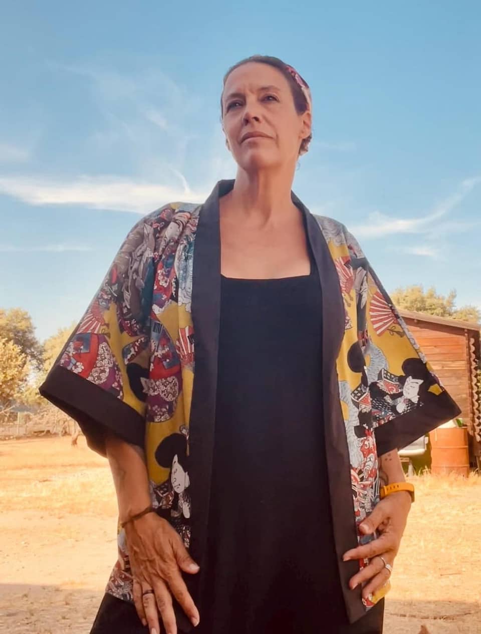 Una chaqueta estilo kimono de color amarillo mostaza con un estampado repetitivo de figuras japonesas tradicionales y abanicos rojos. La prenda tiene mangas anchas y un borde negro sólido a lo largo de la abertura frontal, el cuello y los puños.