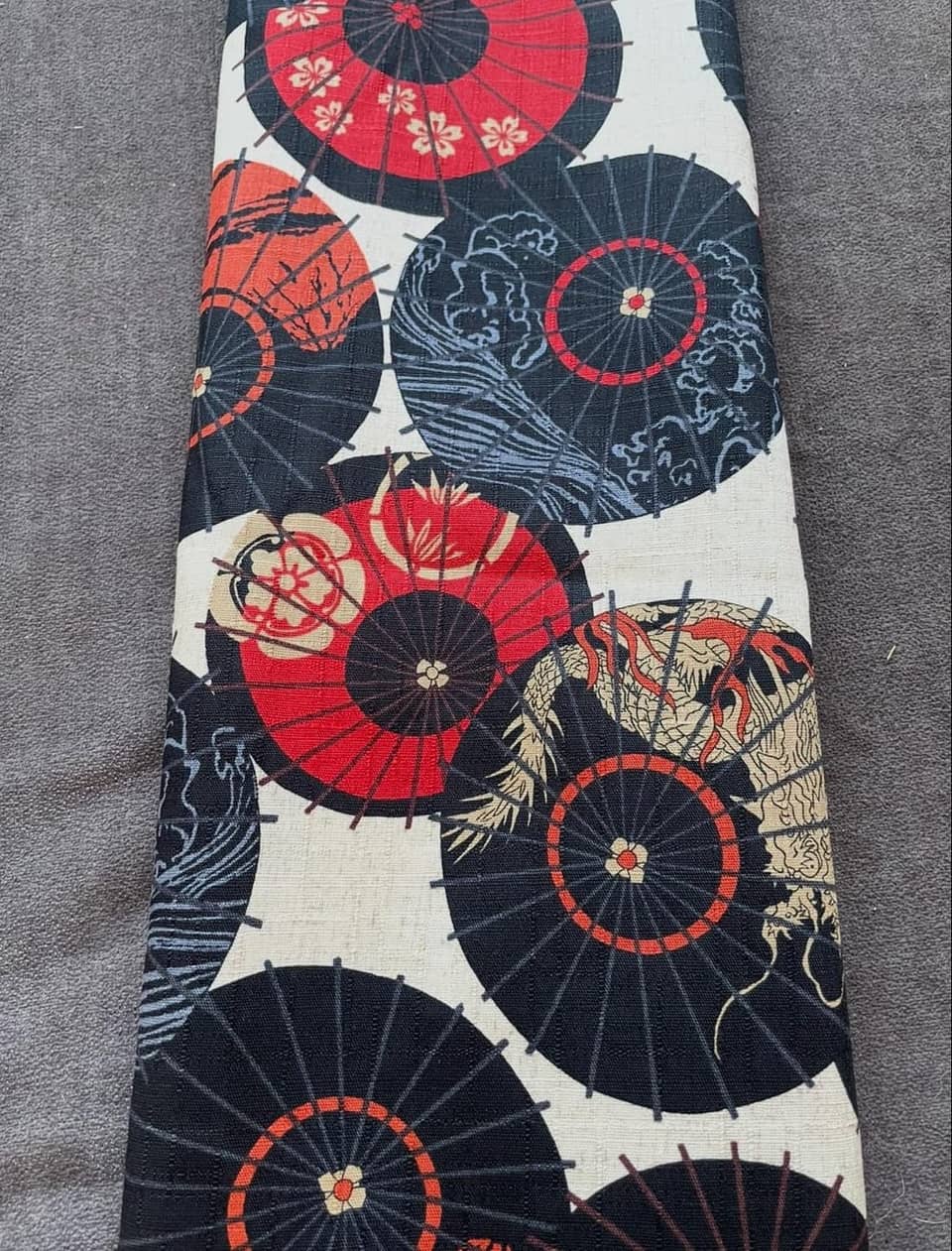 Una pieza de tela rectangular con un patrón repetitivo de sombrillas circulares en negro y rojo. El diseño incluye detalles intrincados como flores de cerezo blancas, motivos de olas azules y una ilustración de un dragón dorado.